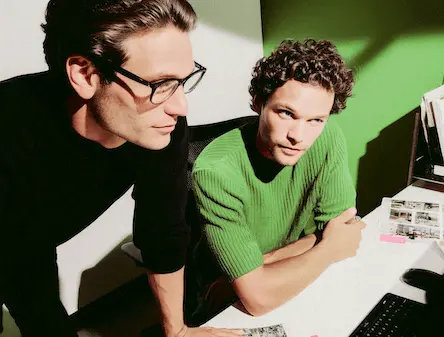 Dois homens olhando para a tela de computador. Um deles veste uma blusa verde e o outro uma blusa preta.